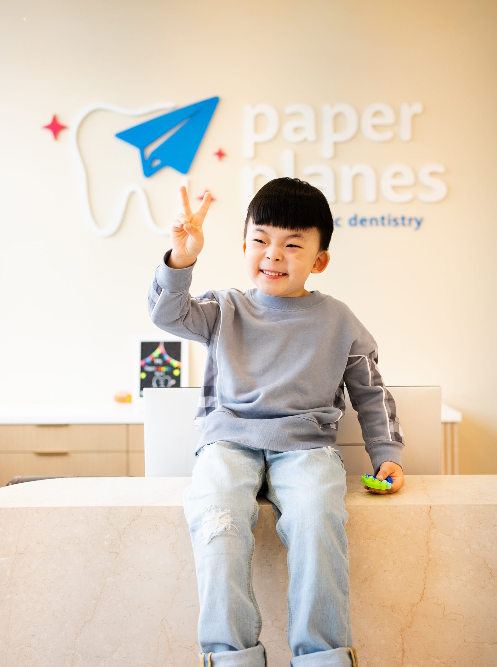 Cheerful young boy making a peace sign while sitting on the reception counter at Paper Planes Pediatric Dentistry.