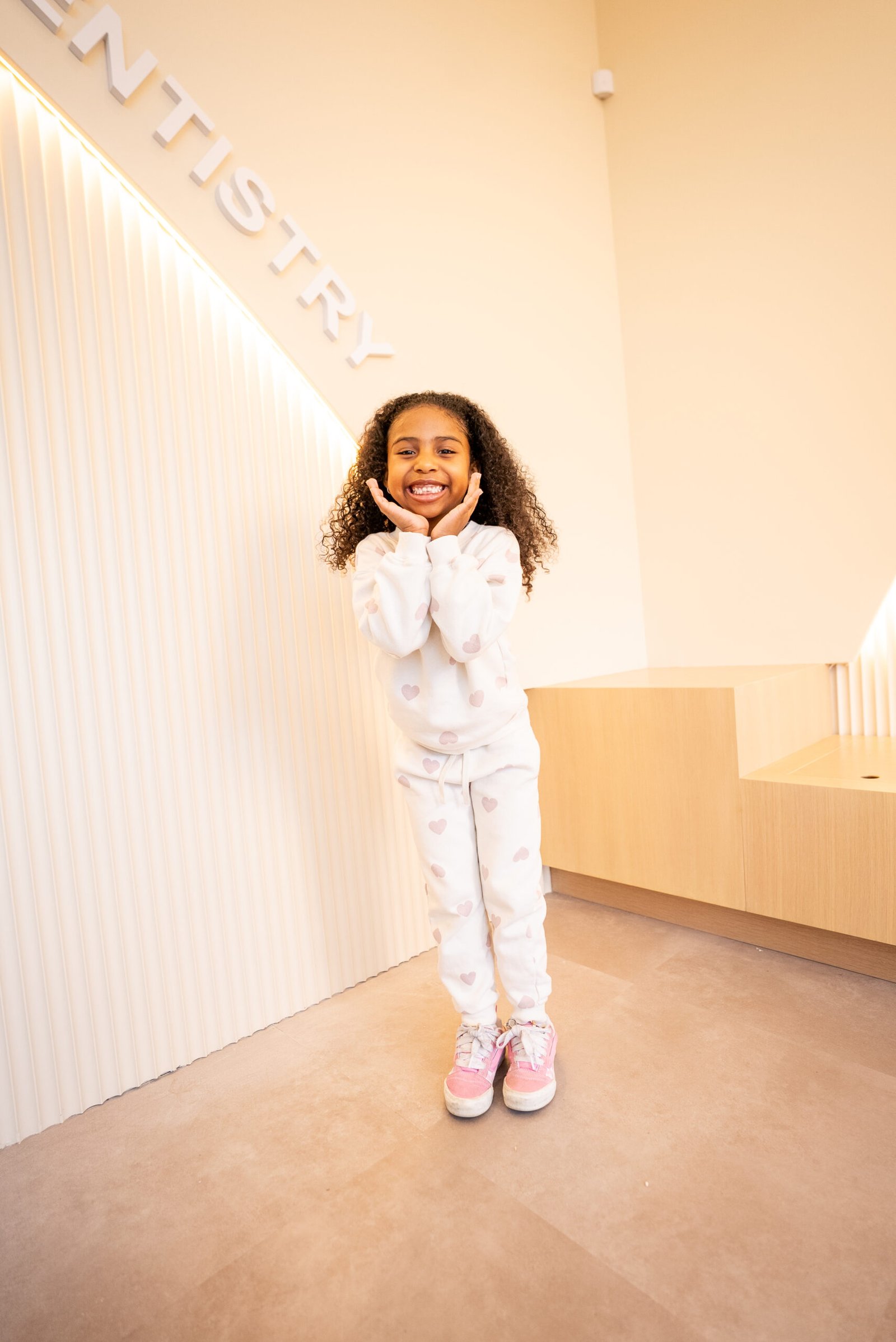 oung girl with curly hair smiling brightly with hands on her cheeks in front of the Paper Planes Pediatric Dentistry wall