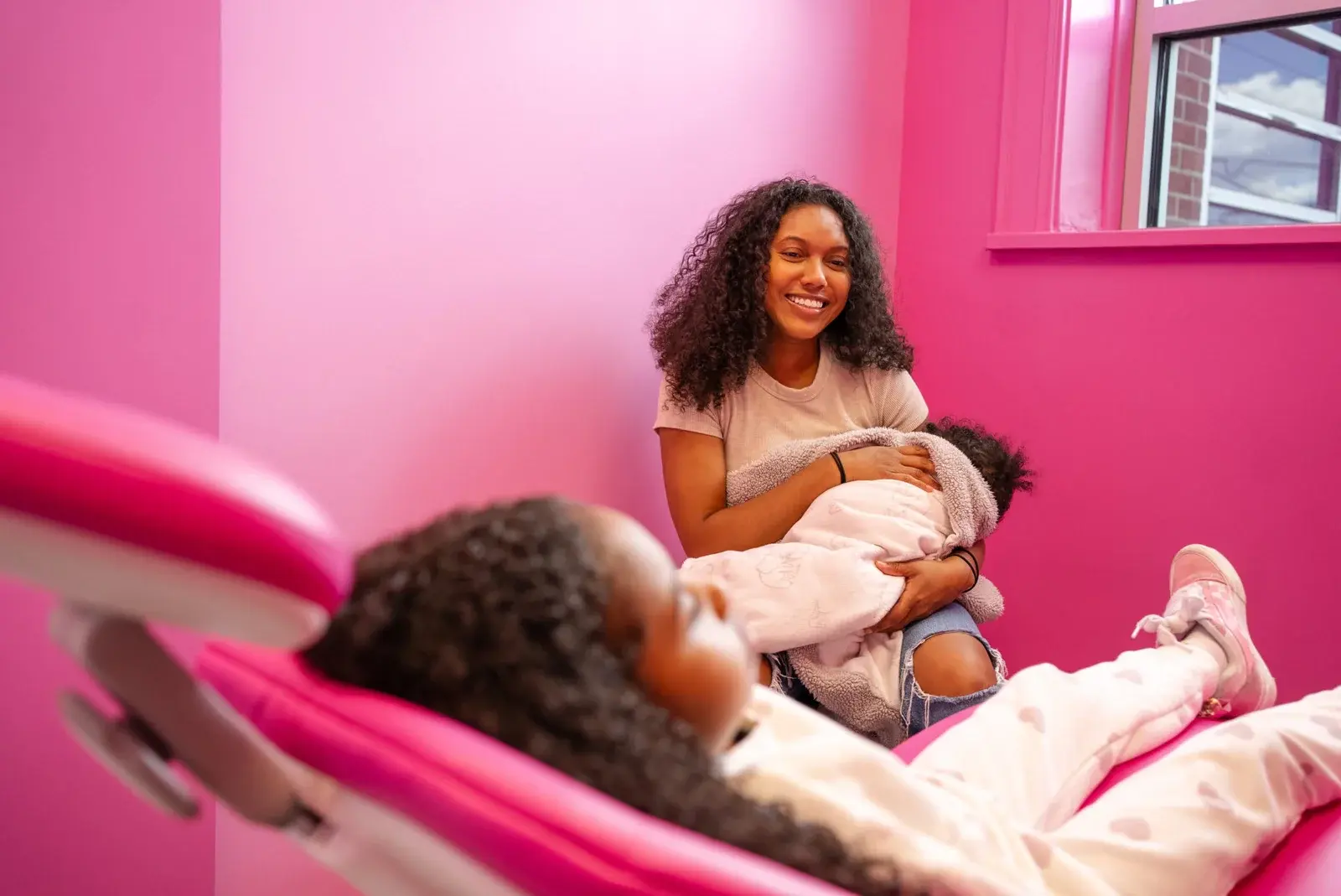 Woman relaxing on couch holding her baby against pink background, conveying comfort, ease, and modern lifestyle.